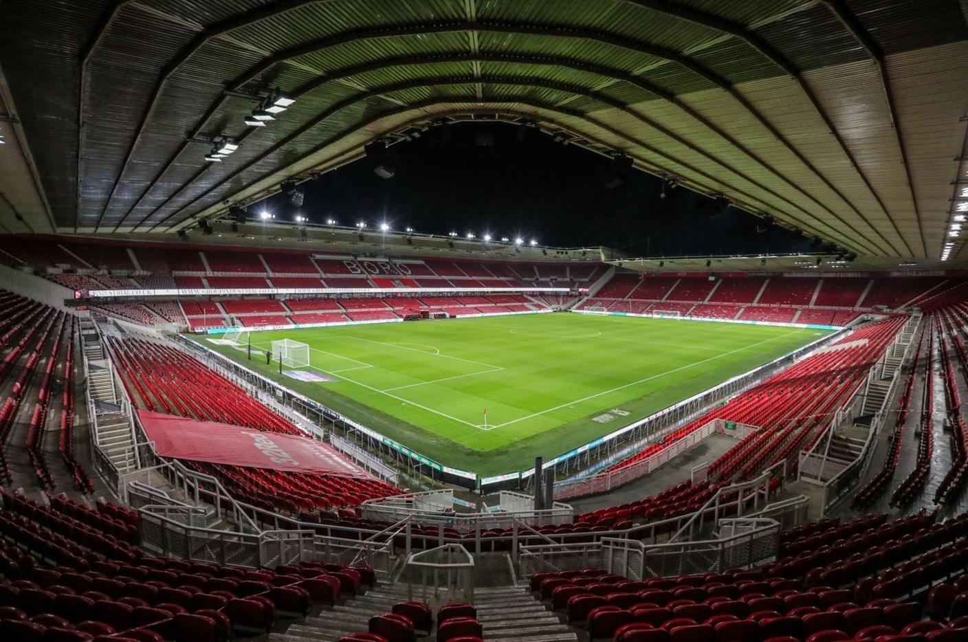 An empty football stadium pictured from a corner high up in a stand. The seats are all red, it is night time and the bright green pitch is lit by floodlights