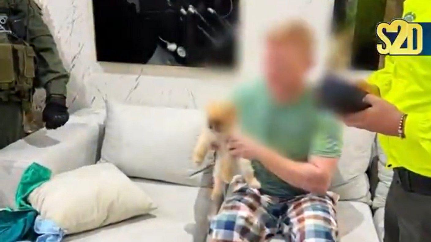 A ginger-haired man wearing a green t-shirt and chequered pyjama bottoms sits on a couch holding a Pomeranian puppy. He is surrounded by police officers. His face has been digitally blurred.