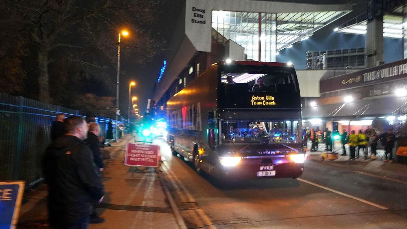 The Aston Villa team bus arrives before the UEFA Europa League match at Villa Park, Birmingham. It is dark outside and the bus has its lights on in front of the lit-up stadium. The sign says Aston Villa Team Coach.