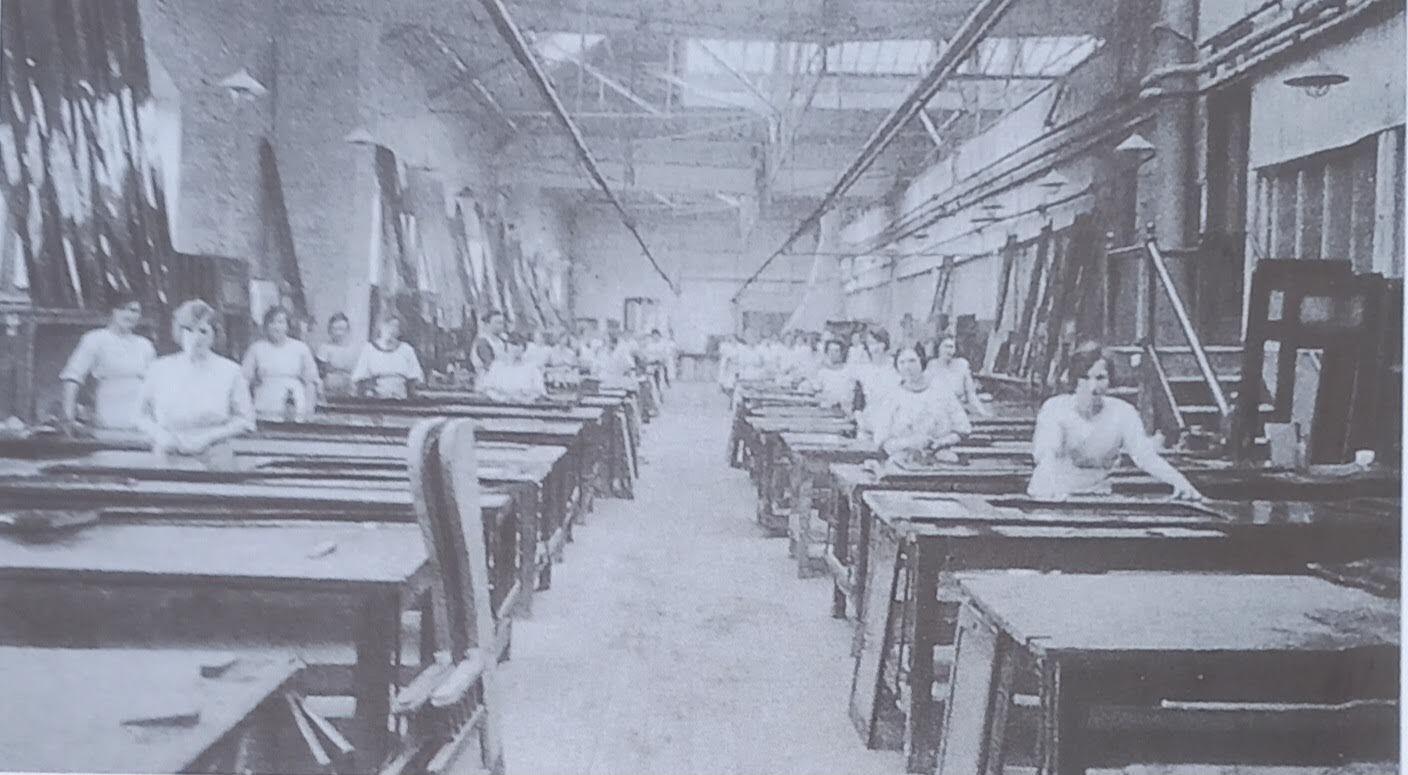 A black and white image of a large workshop with two rows of tables stretching into the distance, spaced closely together with an aisle separating them. A woman stands at every table, facing the camera. One can be seen with her hands resting on a door or window lying on her desk. The ceiling is so high that the roof cannot be seen but there are windows high up on the far wall.  