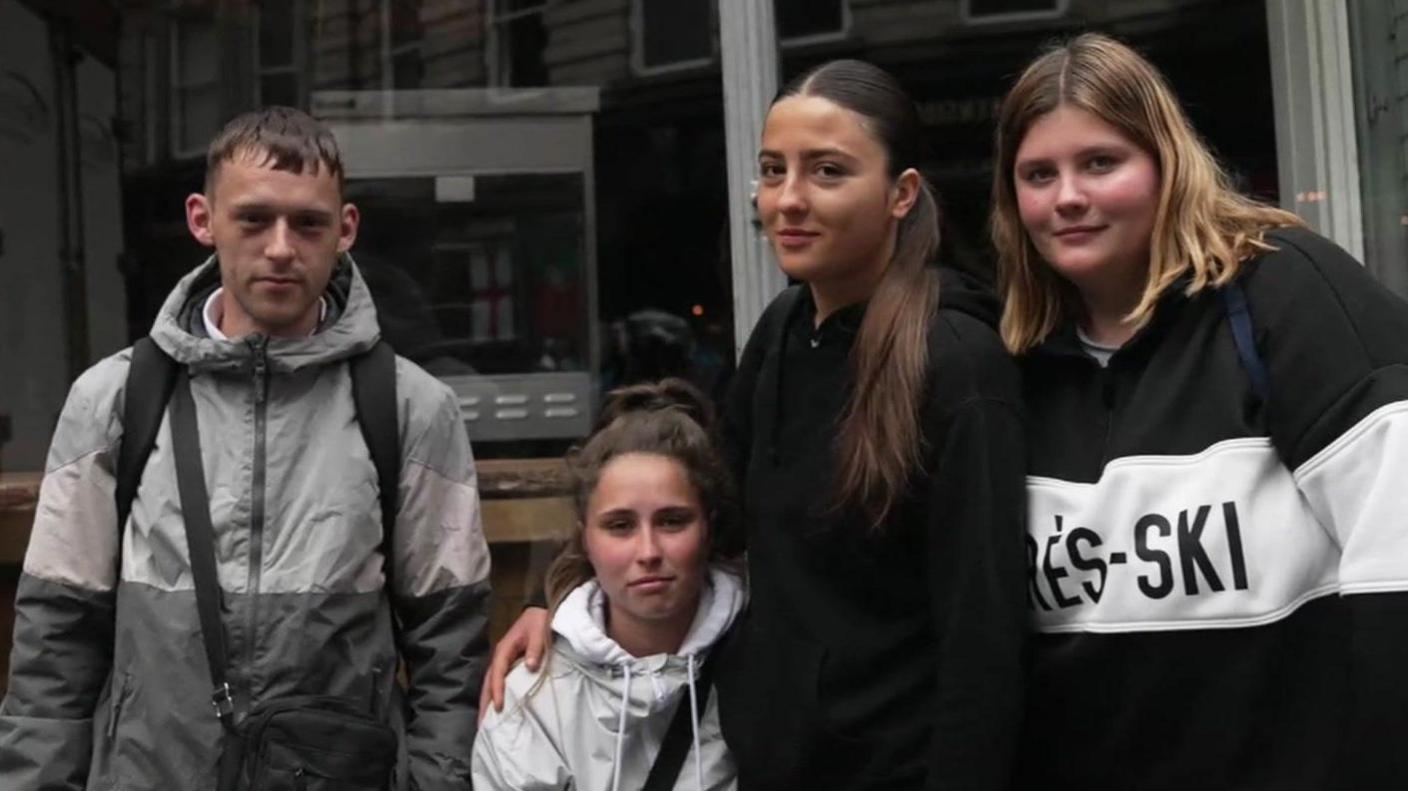 Four young people - three females and one male - standing outside a shop