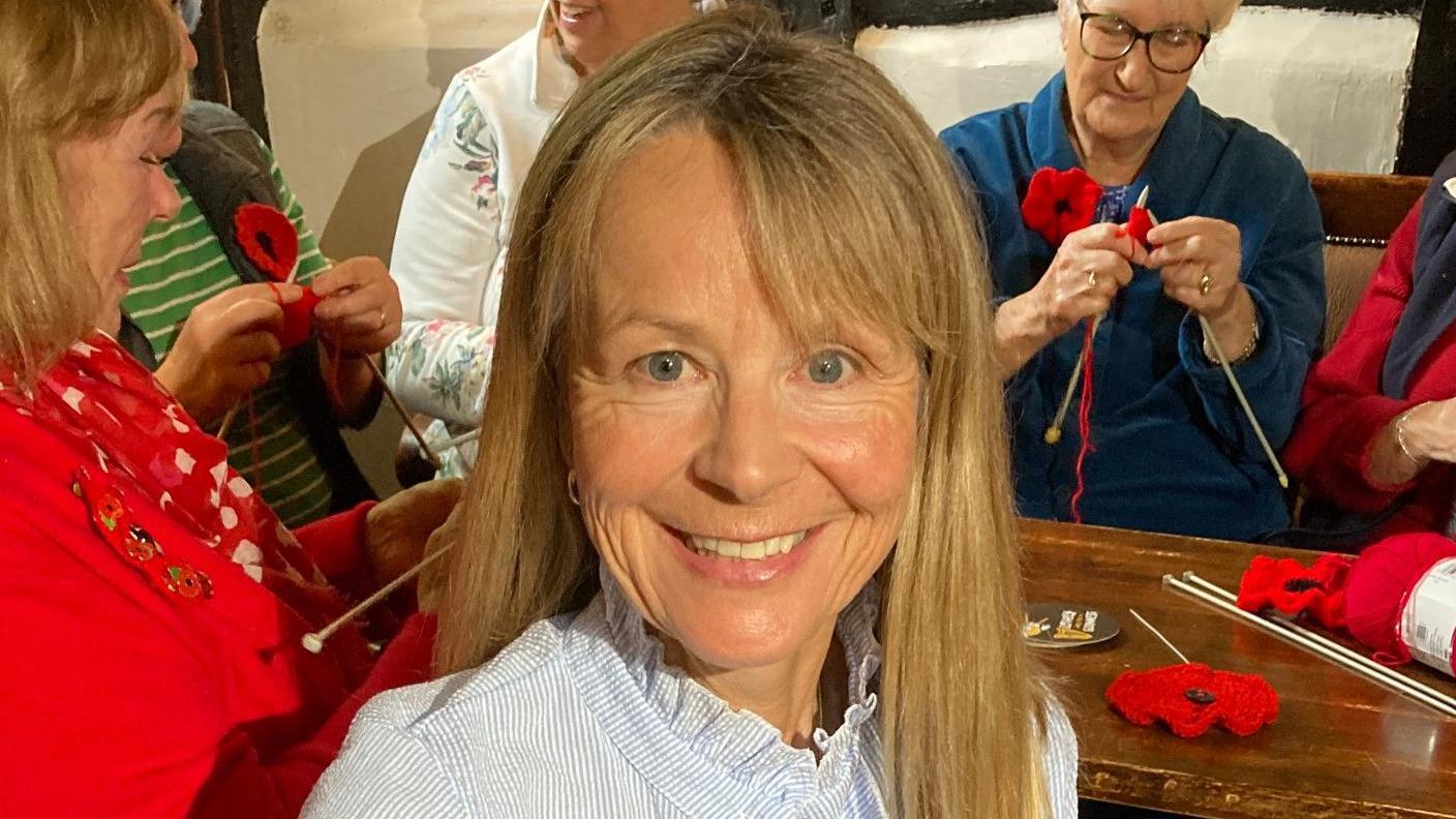 Tina Hill smiling, wearing a blue and white top. She has long hair and other women are behind her knitting by a table that has poppies on.