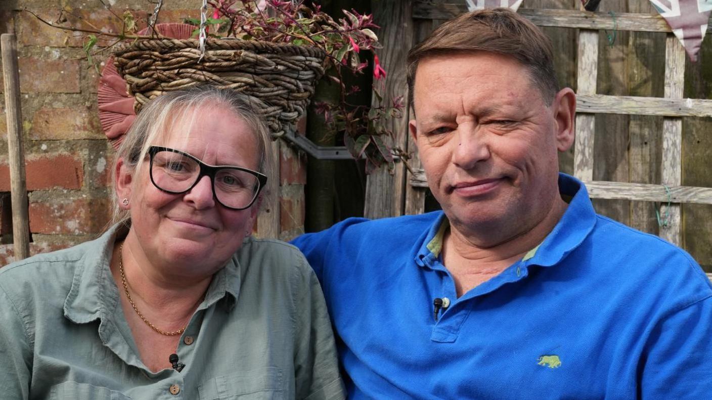 Tony Henderson with his wife Sarah sitting outside their house in Bacton in Norfolk. He is wearing a royal blue polo shirt and he has his arm around his wife's shoulders who is wearing an olive green shirt. 