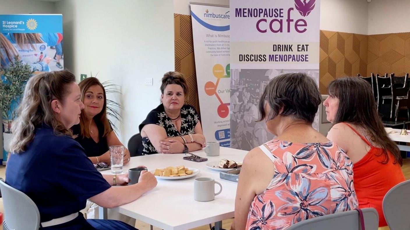 Five women sat around a table with cups of tea, biscuits and cakes. One woman on the left is talking, and the rest are listening.