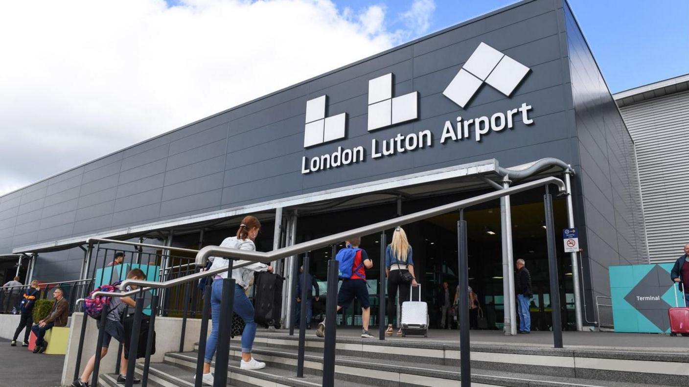 The exterior of London Luton Airport. The branding on the airport building is visible, in white, and in the foreground are steps and some passengers heading to the airport building.