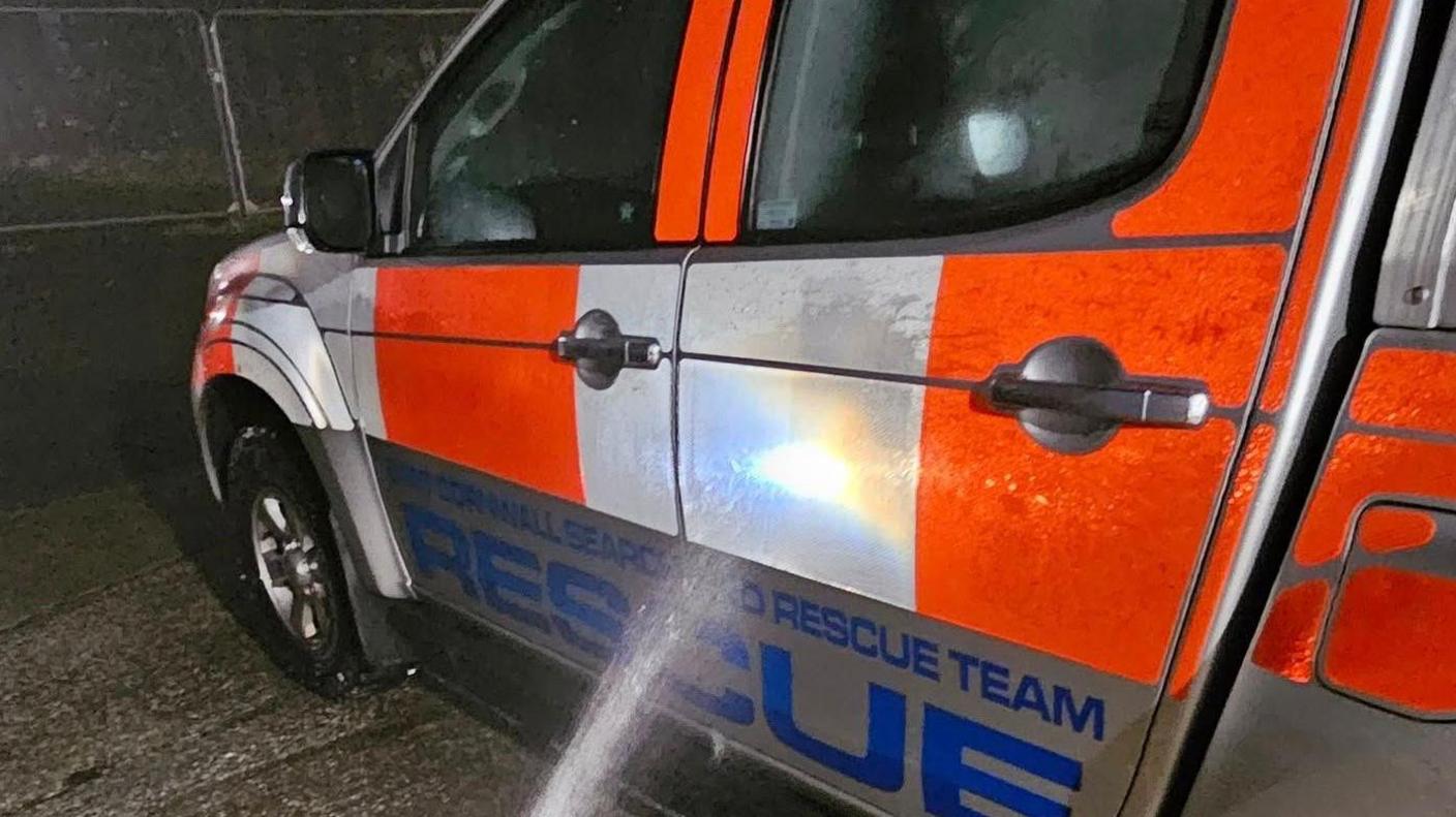 An orange and white van with East Cornwall Search and Rescue written in blue on the side is seen being sprayed down following the operation 