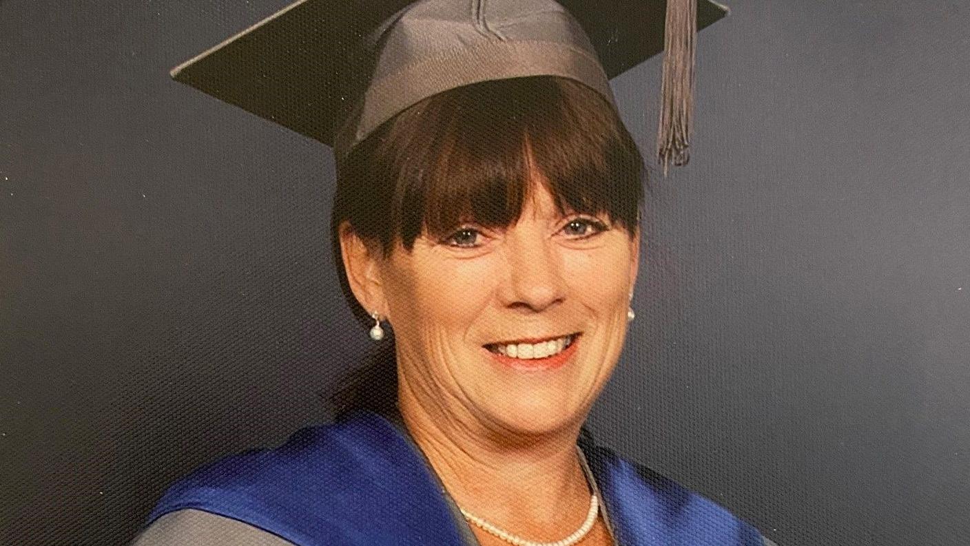 Beverley Pedley is smiling at the camera, wearing a grey mortar board and cloak with a blue sash over the top.