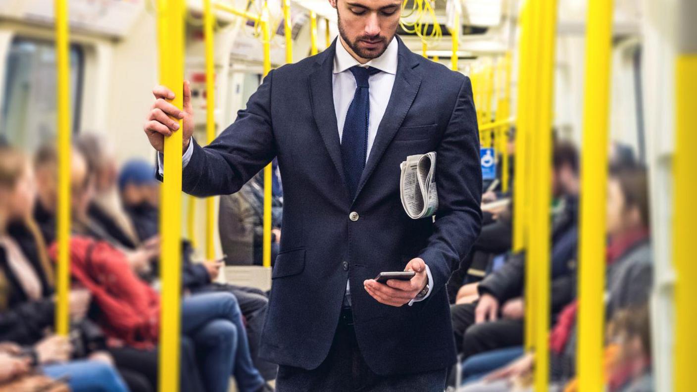 Man in suit standing on Tube train with phone in hand and newspaper under his arm