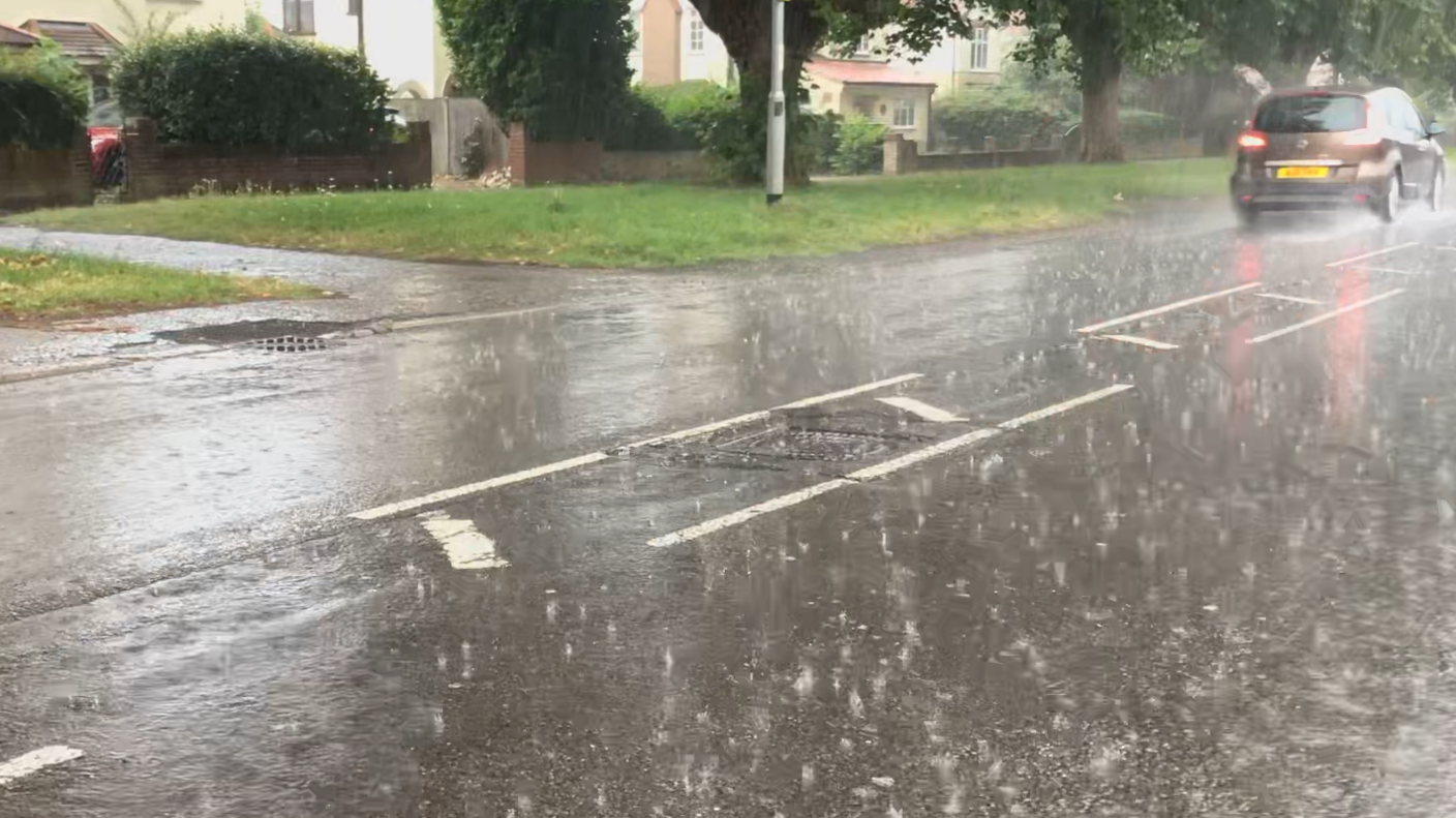 Heavy rain falls on a Norwich road. A car can be seen passing the camera on the road.
