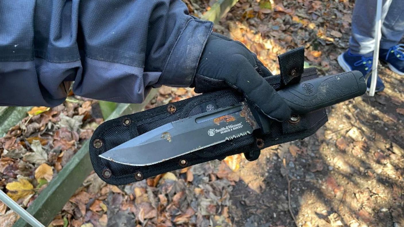 A large survival or hunting-style knife is placed along side the black sheath it was found in. It is rusty and has some mud on the handle.