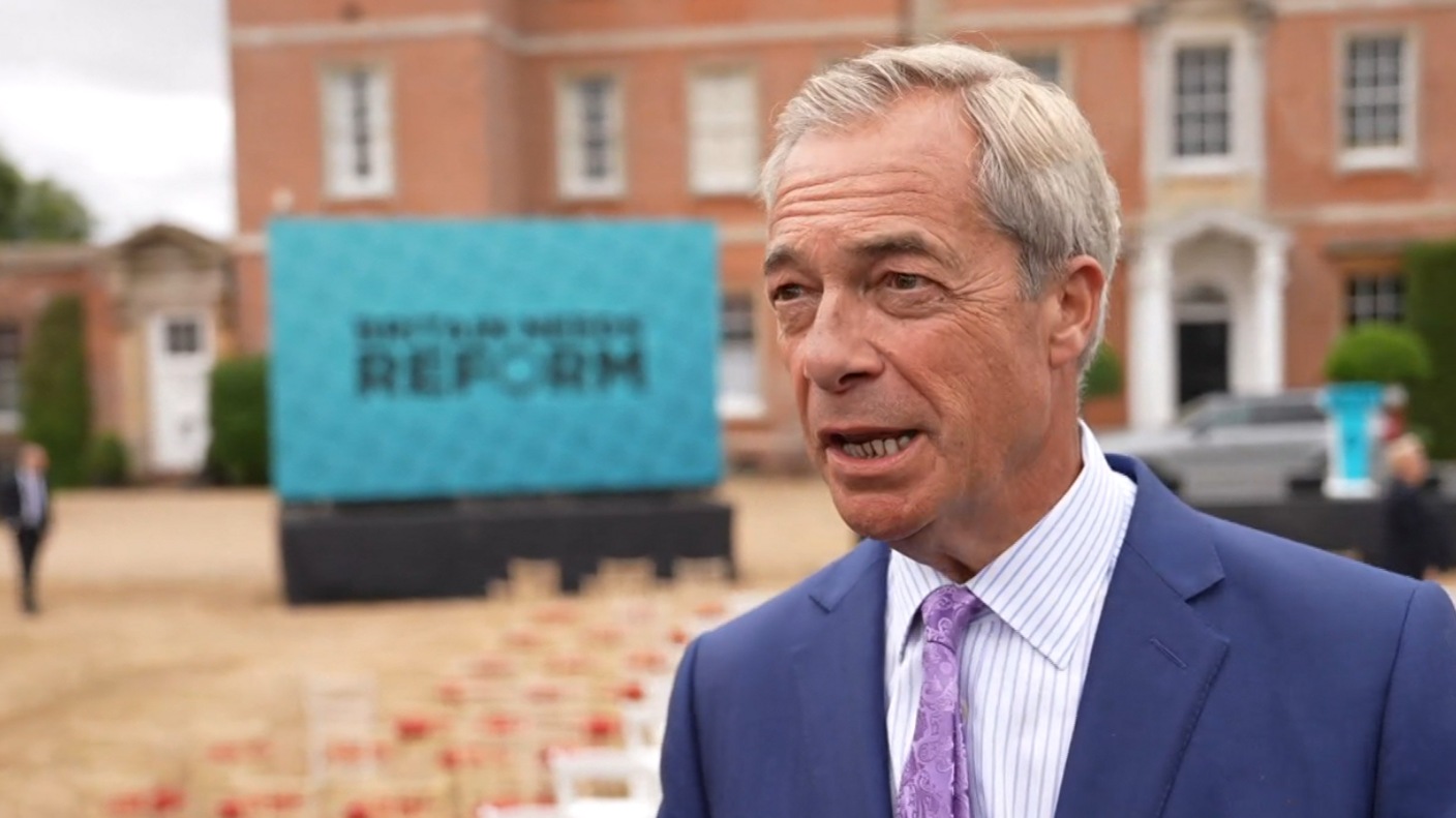 Nigel Farage looks towards a reporter off camera, with his mouth open as he talks, while wearing a blue suit with a striped shirt and purple tie, with chairs outside a stately home blurred in the background