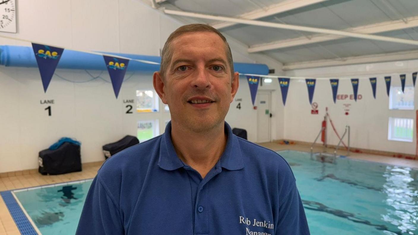 A man is looking at the camera. He's pulling a neutral expression. He's wearing a blue polo shirt. Behind him is a swimming pool, which has bunting hung above it.