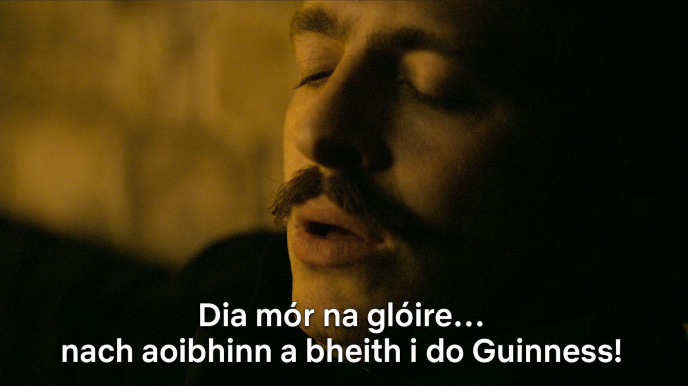 A still from the show, that shows Anthony Boyle as Arthur Guinness with Irish subtitles "Dia mór na glóire ... nach aoibhinn a bheith i do Guinness!"