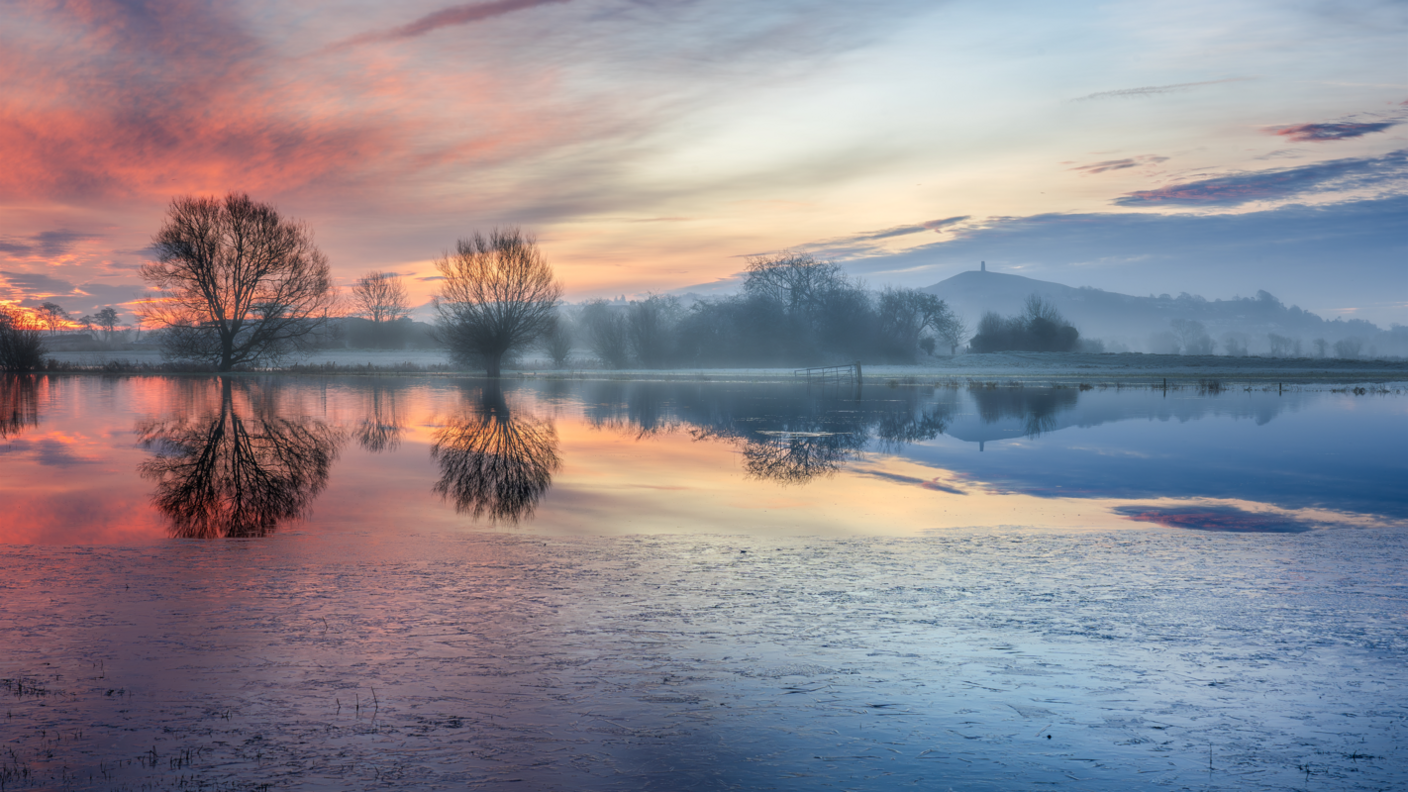 A view of the sunrise over the Somerset Levels. The light is red, orange and blue, the trees are mirrored in the water. In the distance you can see Glastonbury Tor.