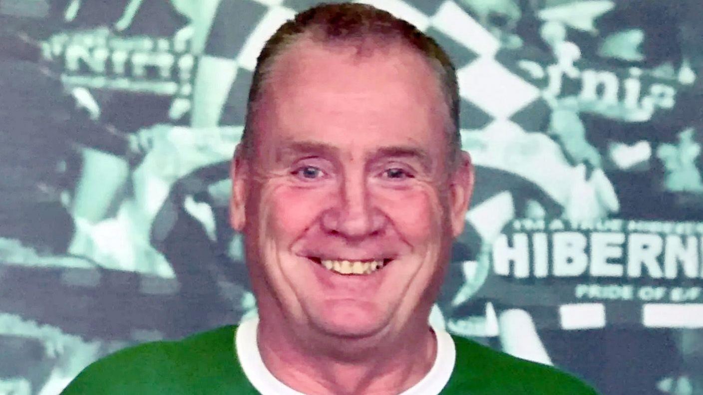 William Budge smiles at the camera. He wears a green and white top. A Hibernian Football Club display is behind him. 