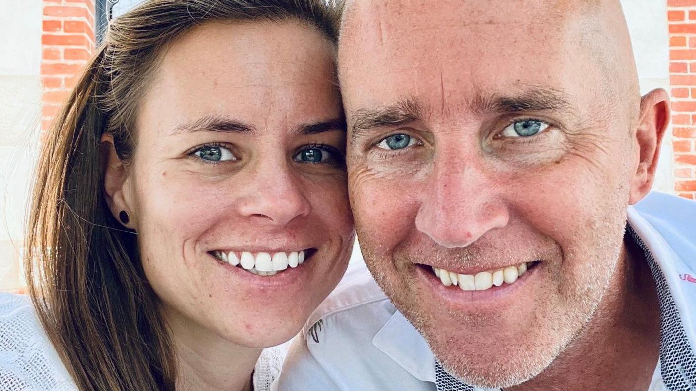 Charlotte Rochenard-Taylor and her husband, Jamie, are next to each other, smiling. A brick building is behind them.