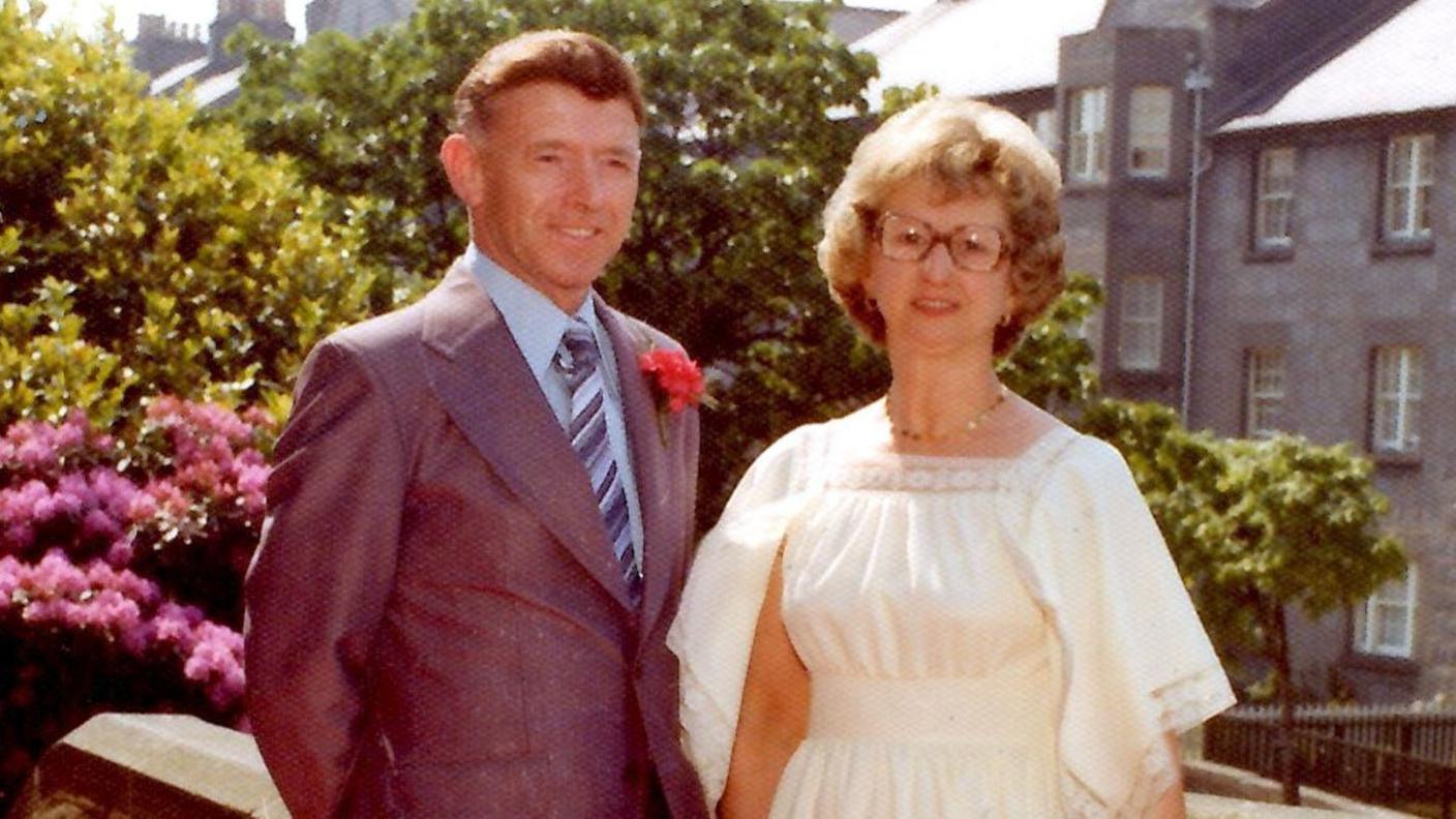 George Murdoch wearing a grey suit, with a light blue shirt and grey and blue striped tie with wife Jessie, wearing a white dress and glasses. The couple are standing in front of plants, one of which has bright pink flowers, and grey tenement blocks in Aberdeen in 1977.