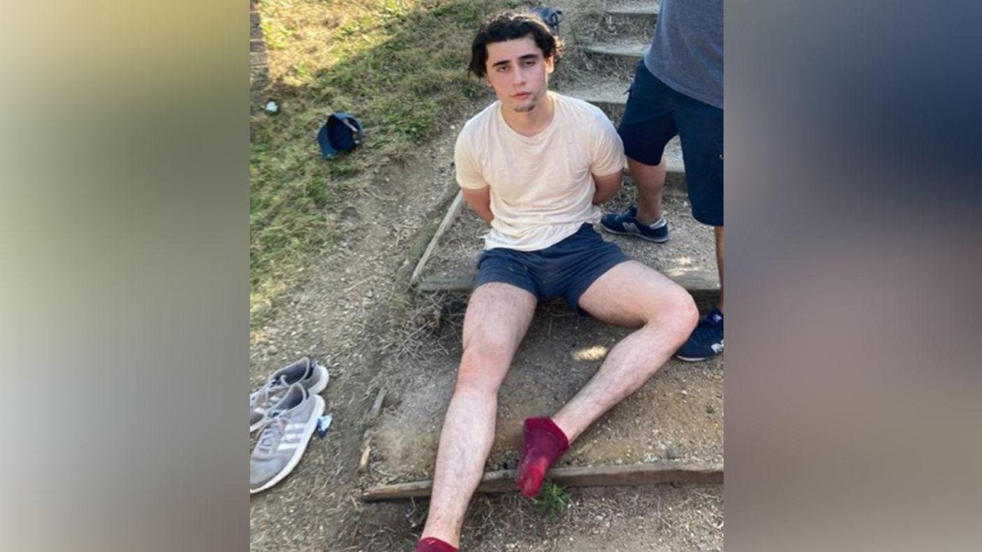 Daniel Khalife sits on the ground with his hands behind his back shortly after his arrest. He is waring a white T-shirt and shorts. 