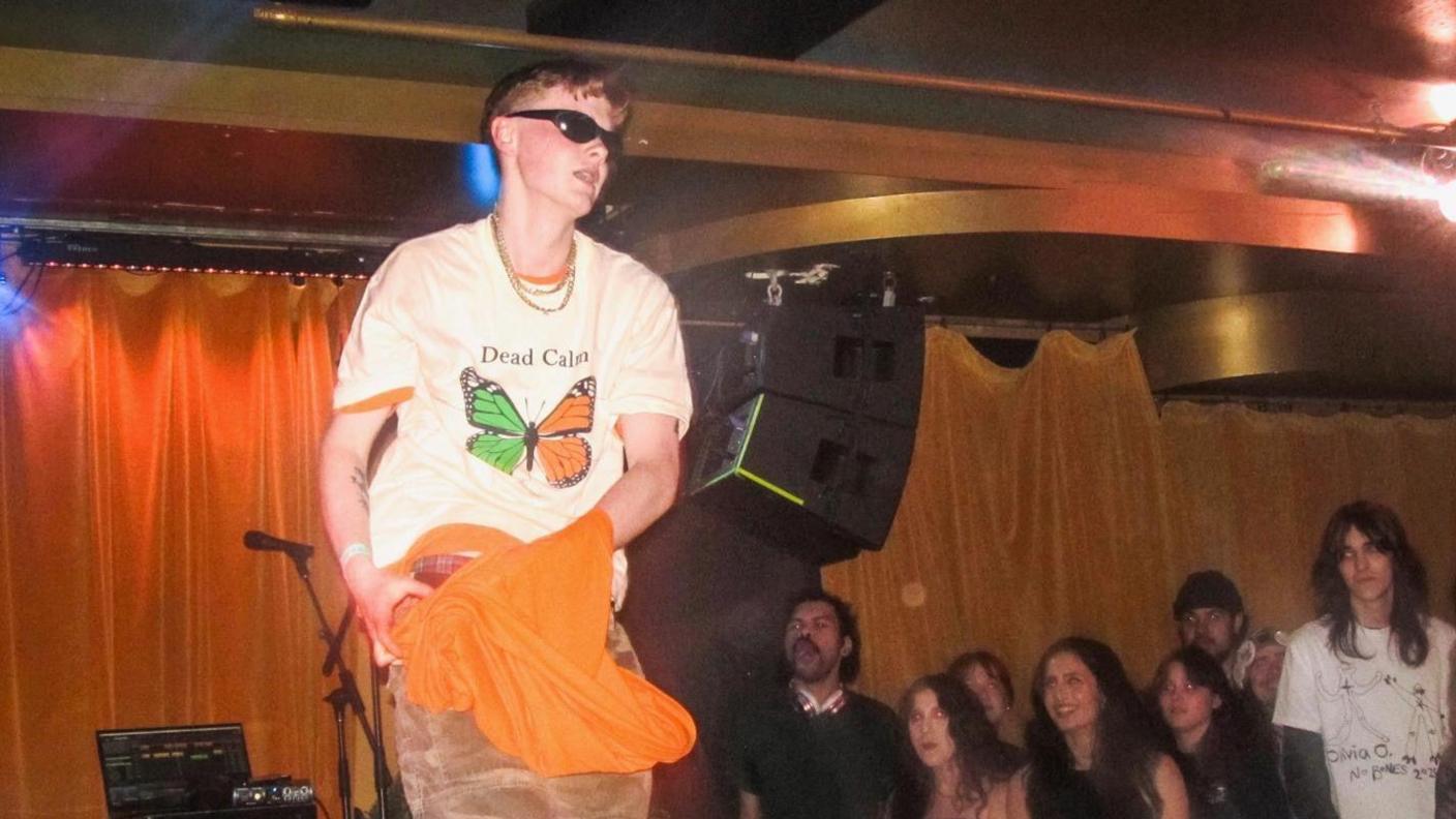 A man in a white top stands on a stage, which is out of frame. There is an orange and green butterfly on his top and he is wearing sunglasses and a gold chain around his neck. A laptop and microphone on a stand can be seen behind him. There is a small crowd of people watching the man perform.