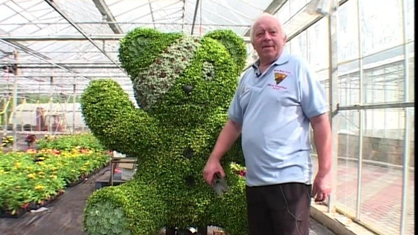 Edwin Smith poses in a greenhouse in front of the giant Pudsey back in 2003. He's wearing a light blue polo shirt and carrying a gardening tool. He has short white hair.  