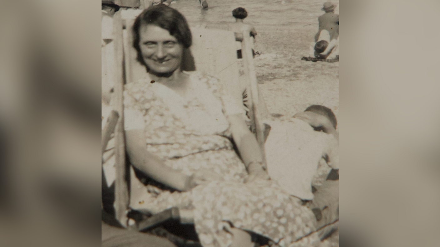 Old photograph of Louisa Dunne. She has dark hair and is sat in a deck chair wearing a floral dress. She is looking at the camera and smiling.