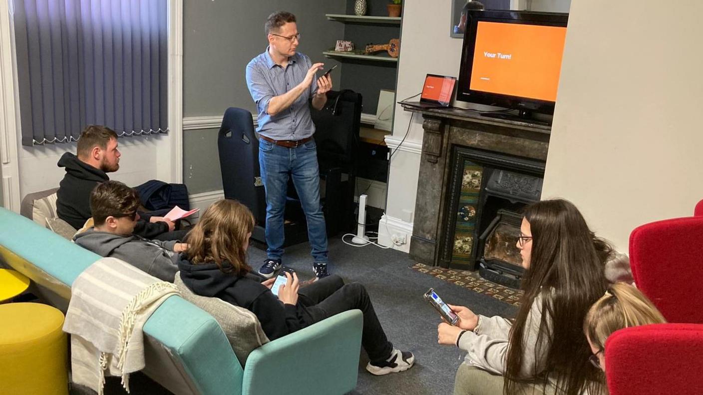 Five young people sat in a room on sofas looking at their phones.  A man stands in the centre with his phone linked to a big screen.