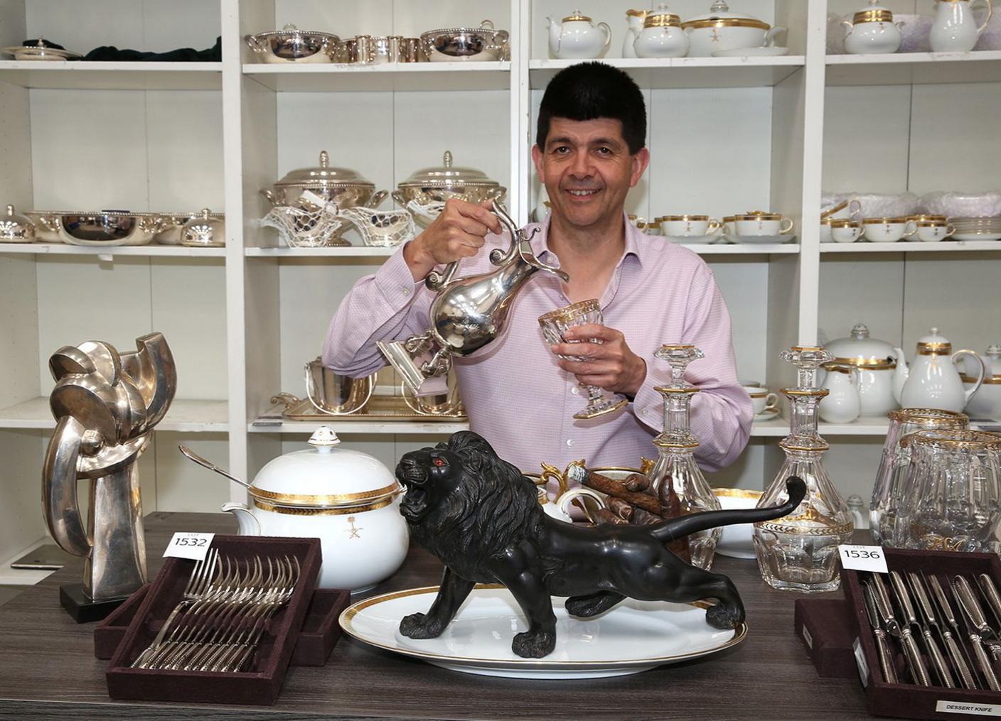 A dark haired man wearing a lilac shirt is standing behind a wooden table. He is pouring water from a silver claret jug into a gold-rimmed glass. On the table are a soup tureen, a black lion sculpture with red eyes, canteens of cutlery and crystal decanters.