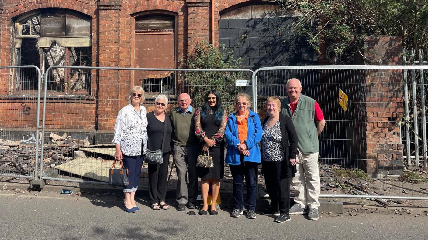 A group of seven people, with a mix of men and women, stand in front of a derelict building on a sunny day. There is grey fencing around the building.