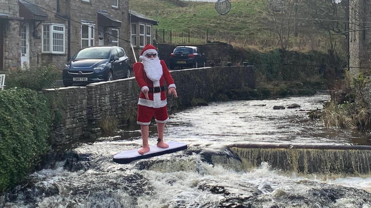 A knitted Father Christmas standing on a surfboard floating over the water on the beck in Hawes. 