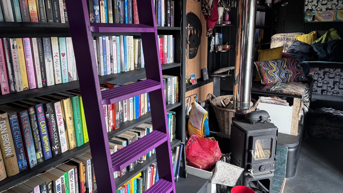 Shelves of books with a purple ladder in front. A black log burner is to the right of it with a metal chimney