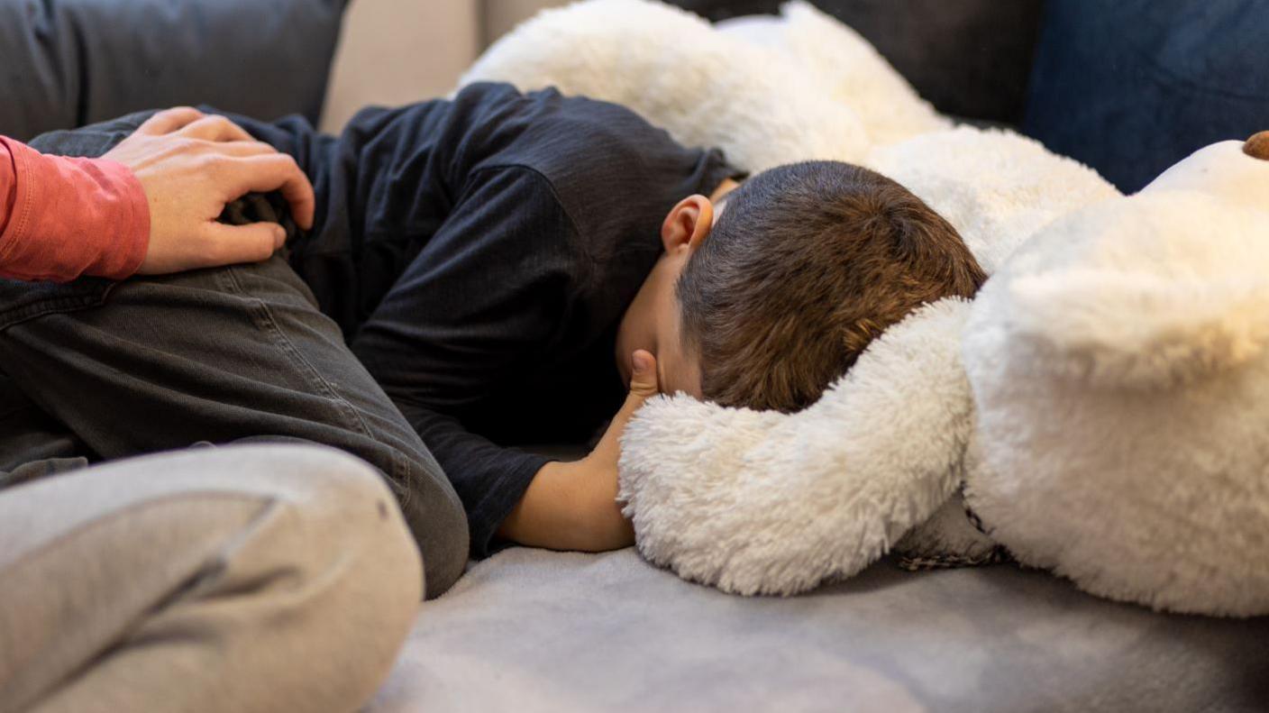 A boy is hiding his face and curled on a sofa. A large teddy bear is next to him. Someone's hand is comforting him.