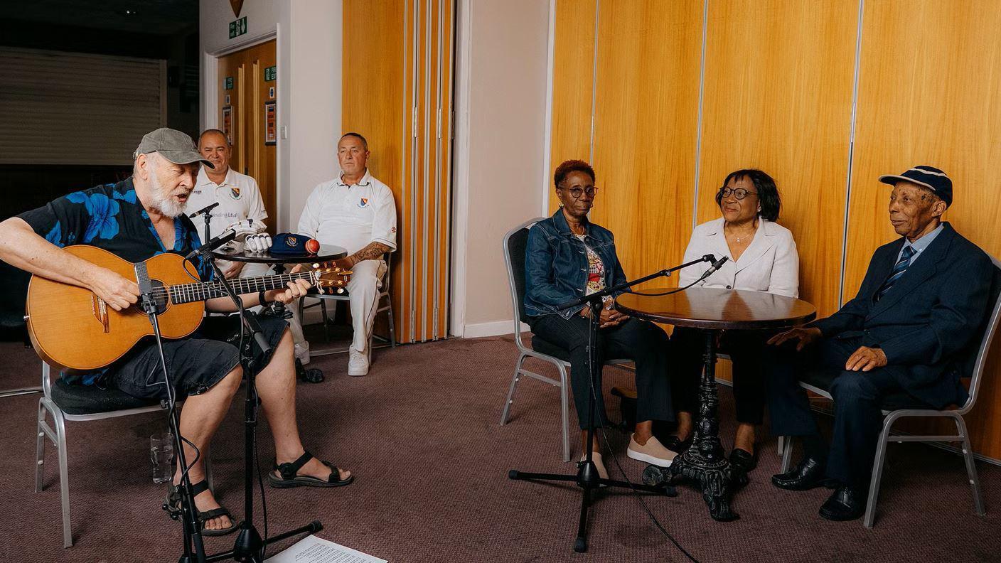 Richard Thompson playing a guitar and singing as Siggy Cragwell sits to one side at a table with two women. There are two cricketers in cricket attire in the background.