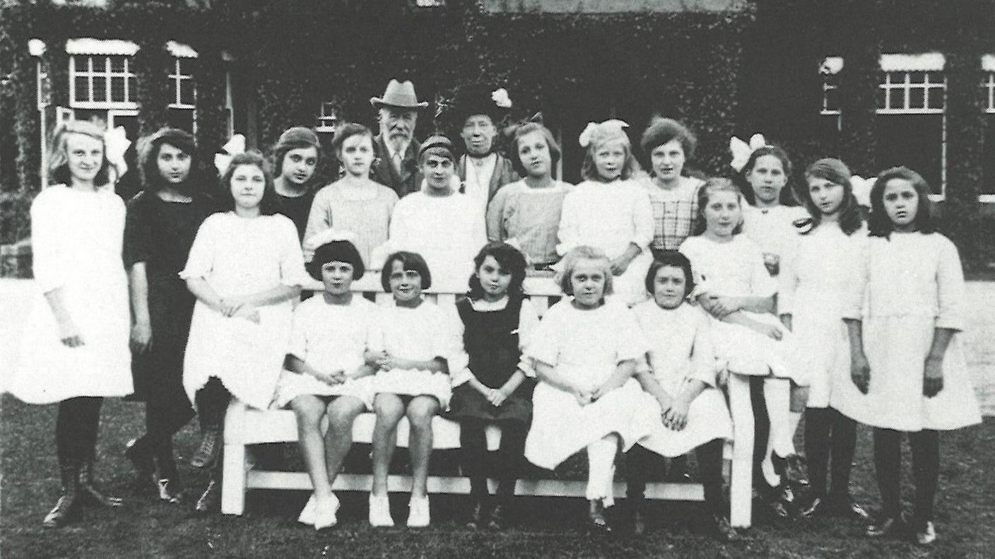 A black and white photo of a man wearing a light hat and a woman wearing a black hat. They are pictured with  girls, who are mainly wearing white dresses. 