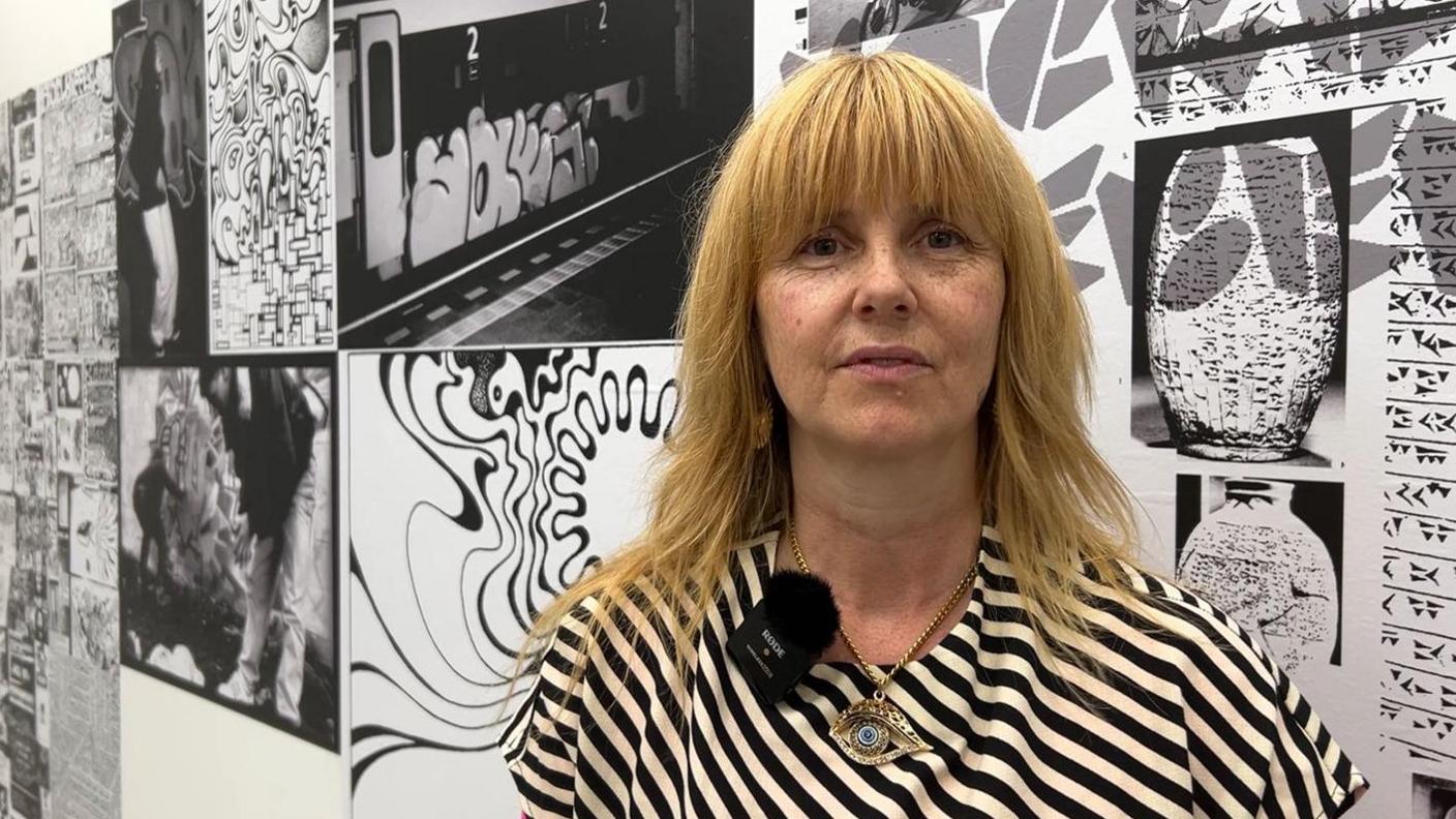 Lonny Pop has strawberry-blonde hair and is wearing a stripey monochrome top. She is standing in front of a montage graffiti wall