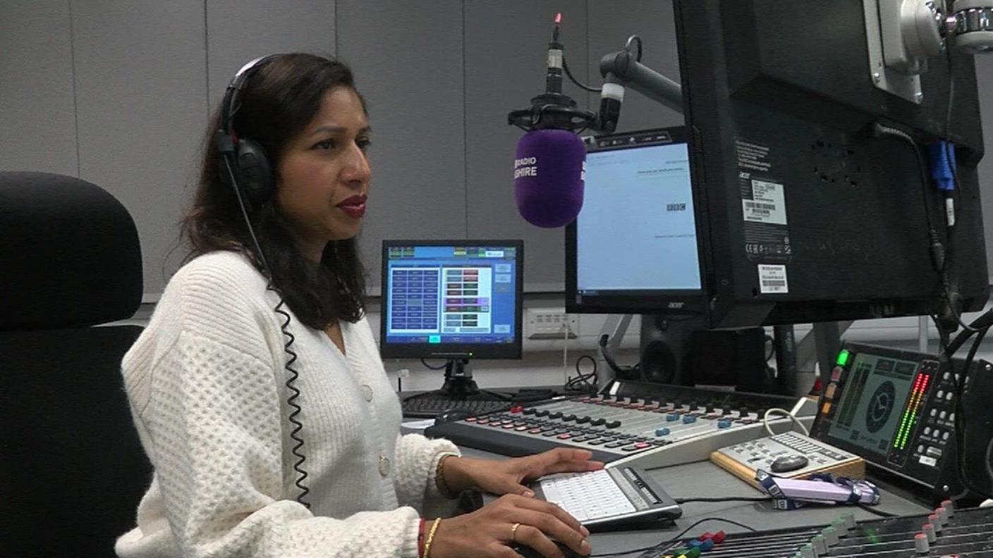 Rena Annobil sitting in the radio studio. She has long dark hair and is wearing a white jumper.