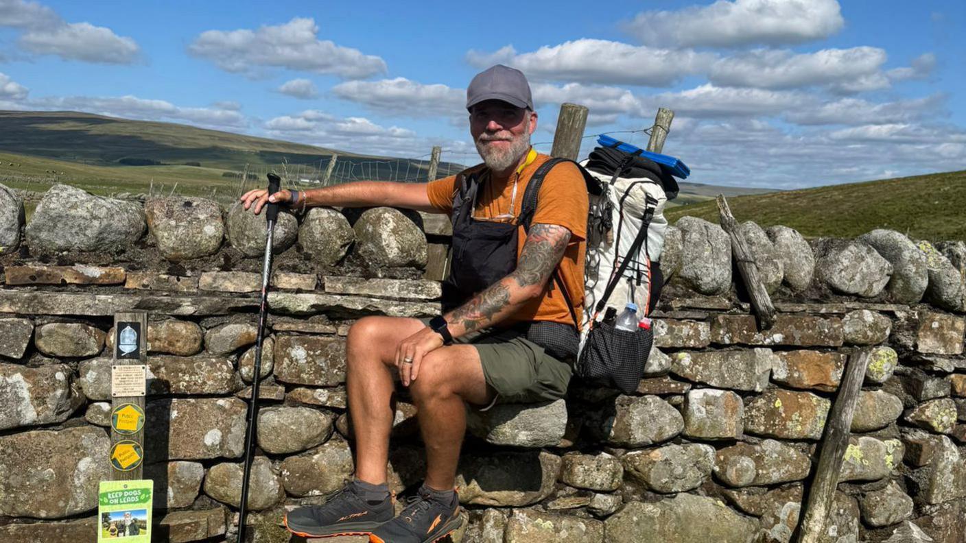 Mr Neal sits steps at the side of a stone wall. He wears a cap, orange T-shirt, green shorts, hiking boots and a walking stick rests in his hand. He has a large pack resting on his back.