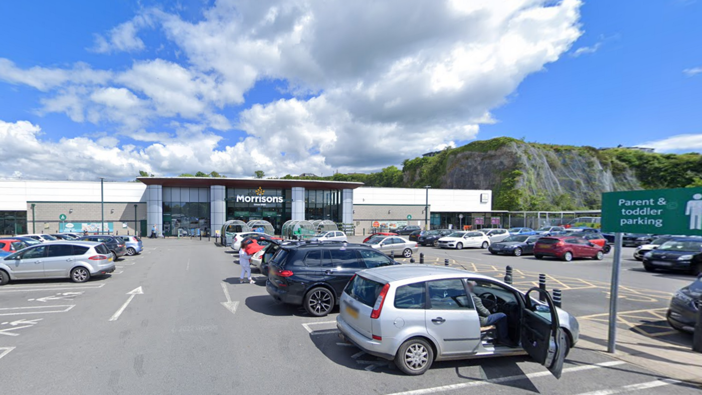 Looking towards the glass-fronted Morrisons supermarket from the car park on a bright day. There are parked cars in the foreground, including one with its driver door open, and a sign indicating parking for parents with toddlers. 