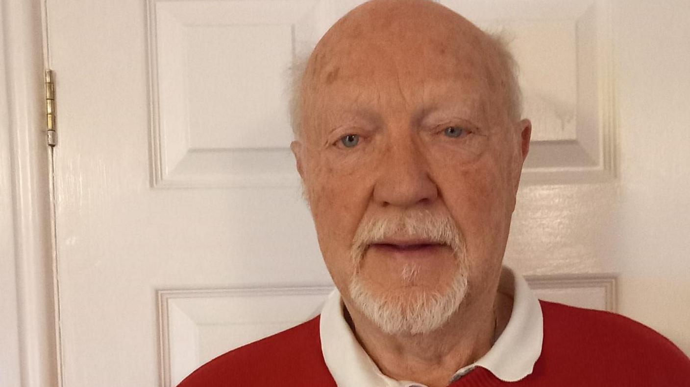 John Irvine is wearing a red jumper with a white collar. He has a white goatee and white hair.