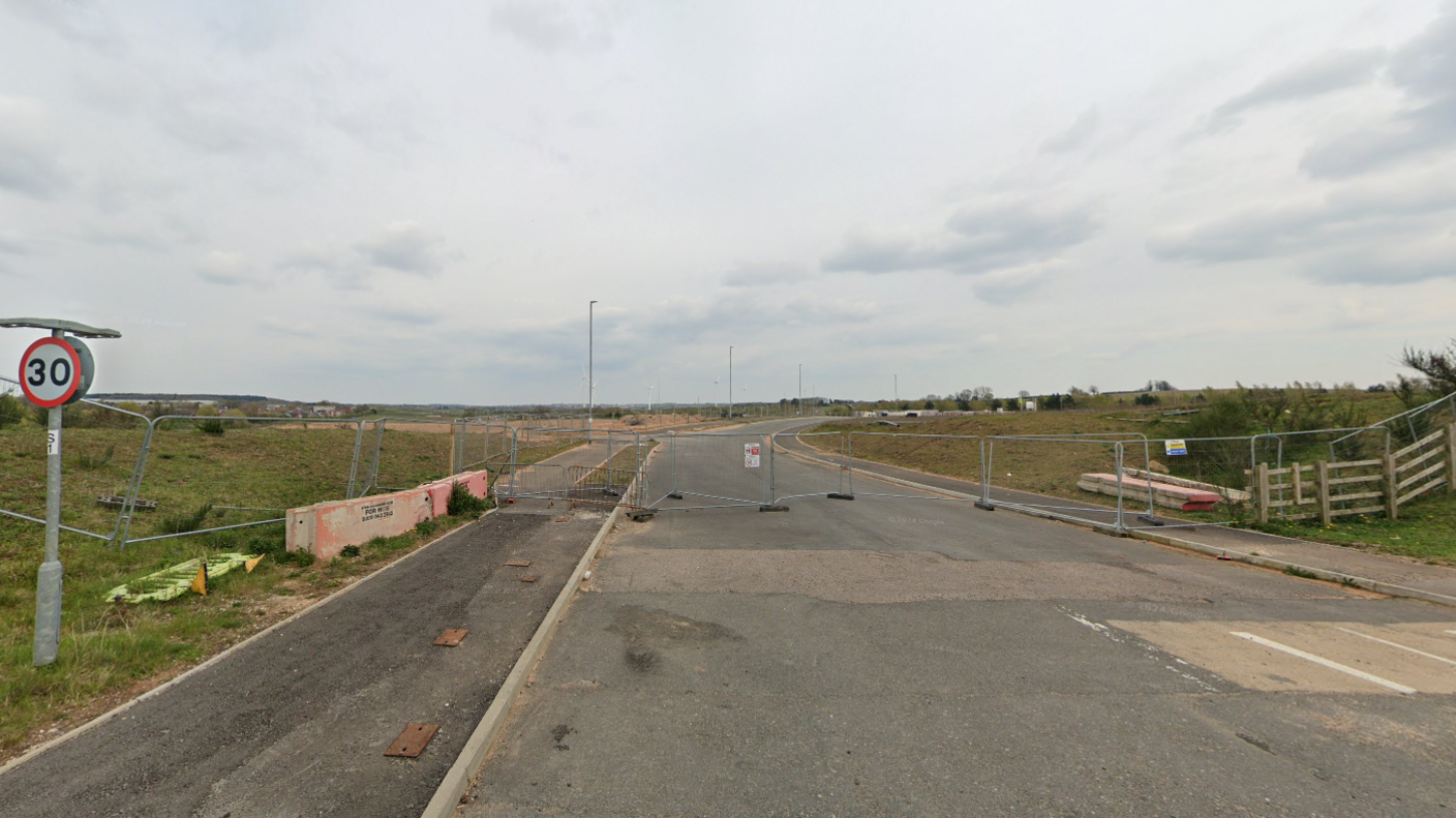 A fence across a road leading in to a green space beyond it