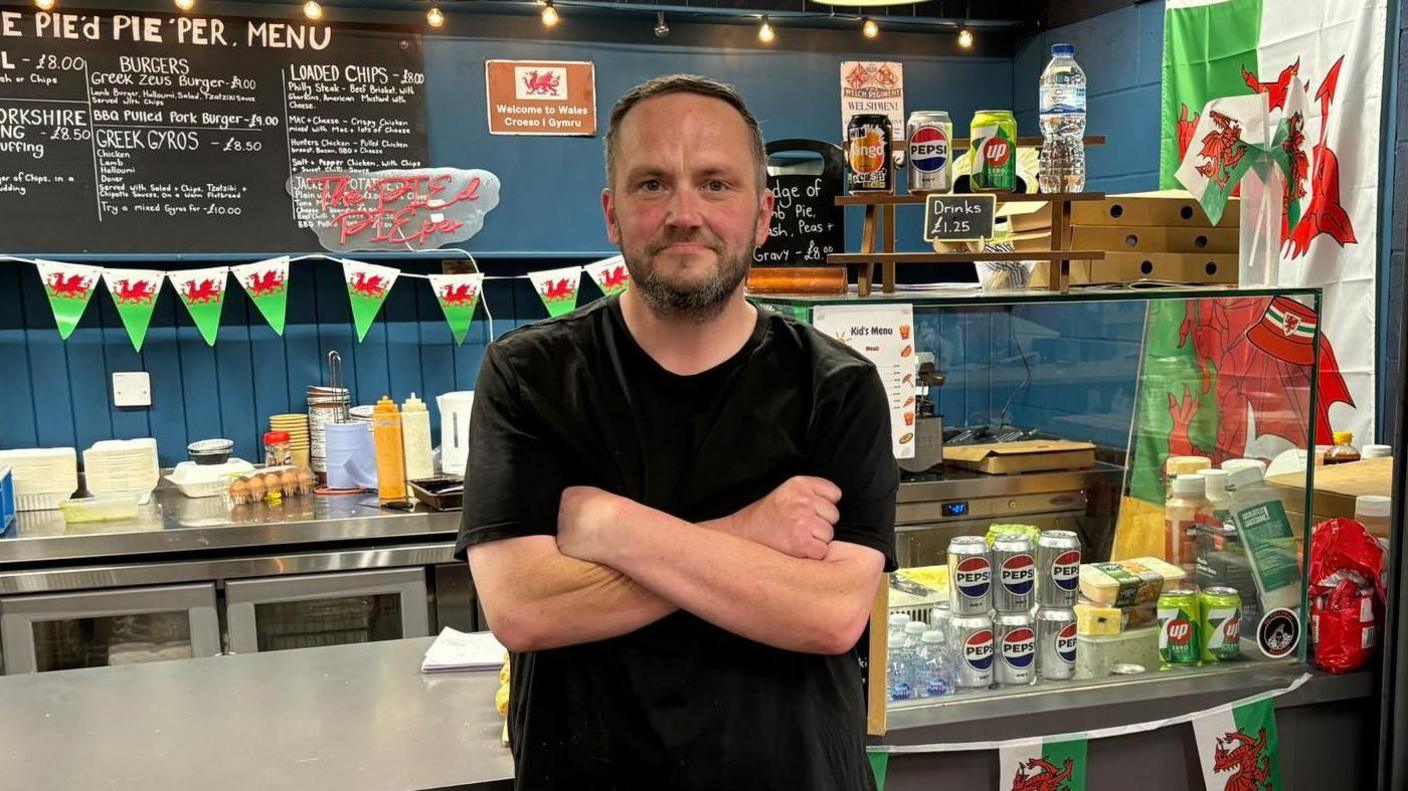 A man in a black t-shirt in a shop stood with his arms folded. There is a pie menu on a blackboard behind him