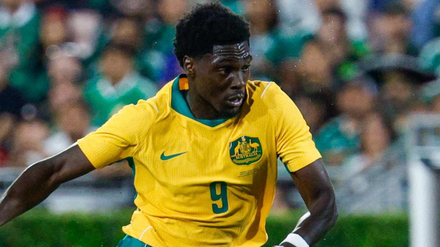 Mohamed Toure runs during a game while wearing Australia's yellow shirt with green trim on the neck, a green Nike tick and Australia badge.