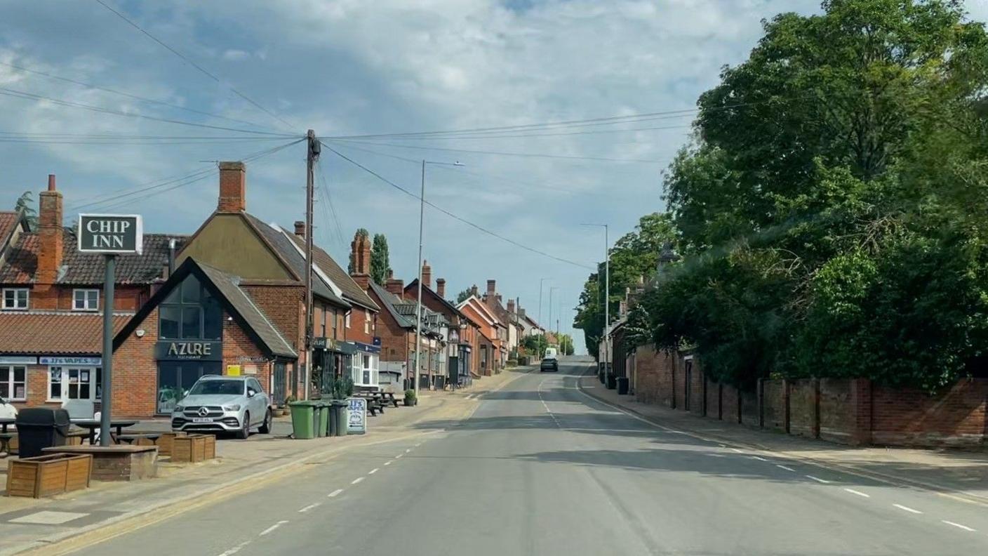 A road with only one car on it is shown with a fish and chip shop at the front of a row of shops on the left and trees on the right.