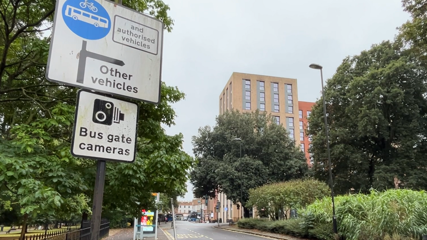 St Vincent's bus gate, with a sign in the foreground warning of the road restriction and the enforcement camera. A bus stop is situated in the road, between a park and a smaller green area.