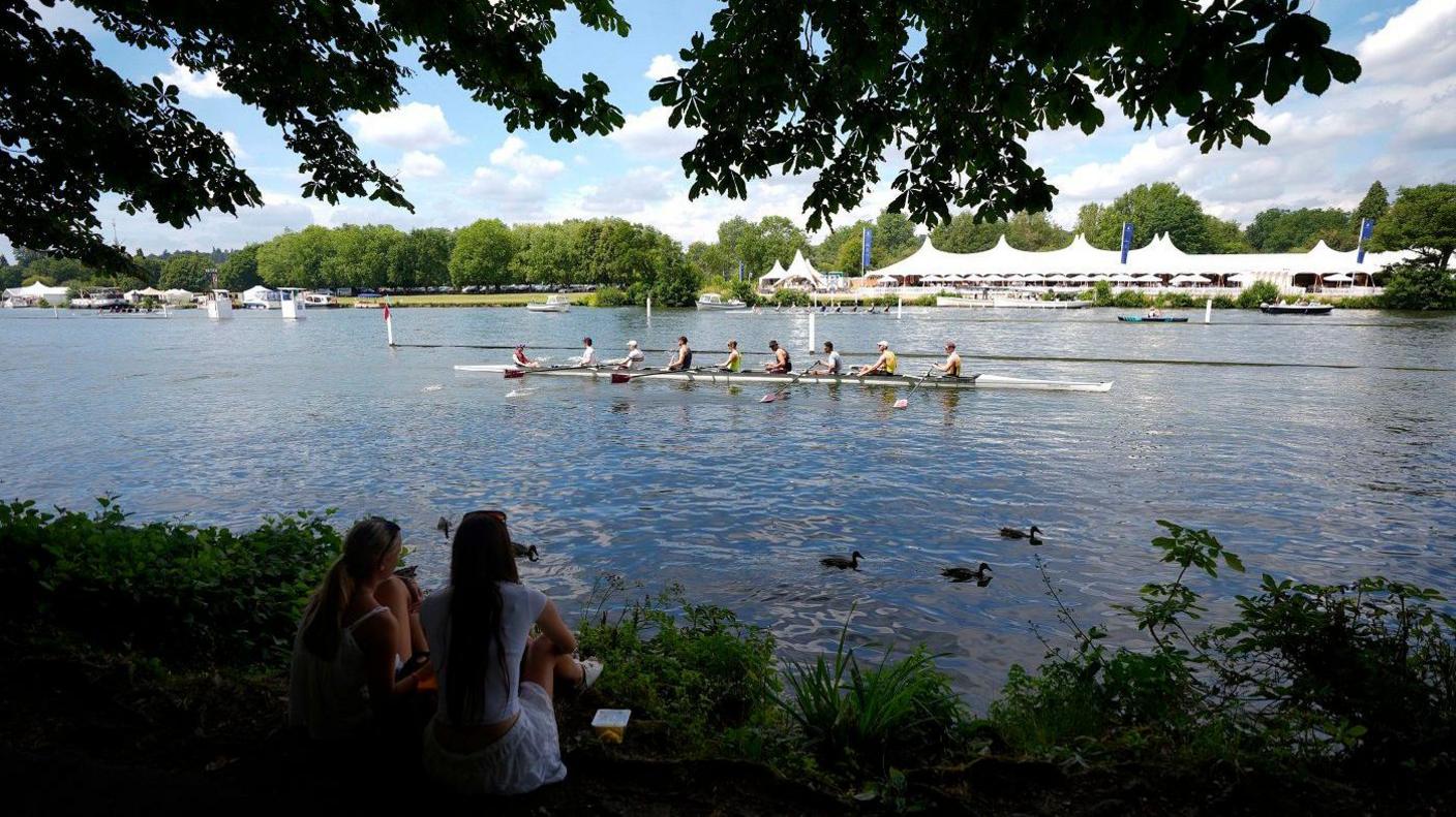 Two women sitting looking out over the river as a rowing boat goes past. There's a row of white tents across the water.