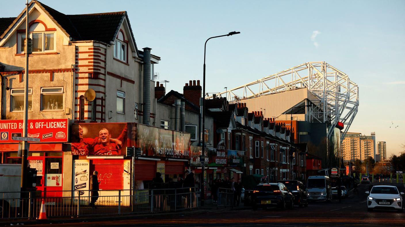 A Manchester United-themed off-licence can be seen in front of a row of terraced homes that open out alongside a road towards a stand of Old Trafford.