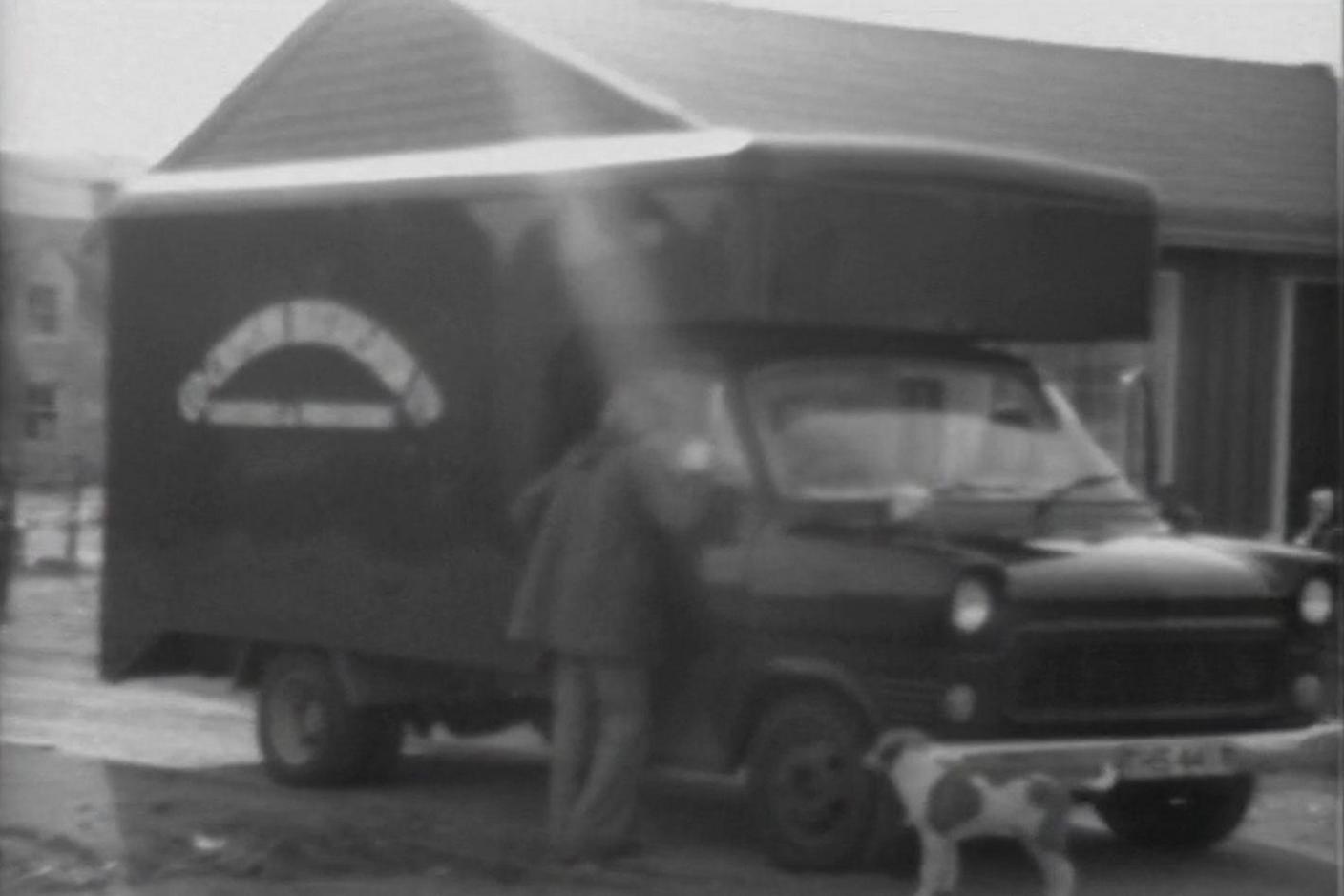 A man and his small dog are looking at a Ford Transit van outside a wooden building. The low quality of the pictures and the sun flare makes the name written on the side of the van hard to read.