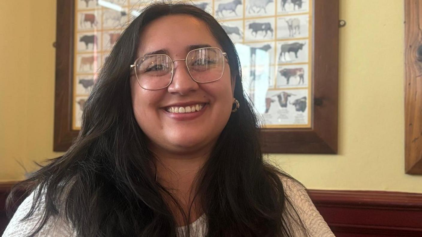 Natasha Cham smiling into the camera. She has dark, long hair and is wearing large silver glasses. She is standing in front of a framed photo of cows.