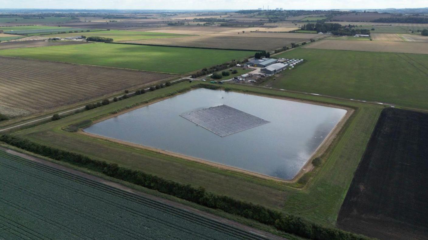 A man-made farm reservoir stands in the middle of green fields. In the middle of the reservoir there are floating solar panels.