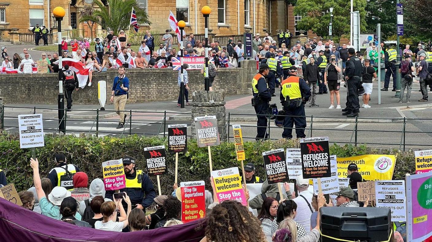 Two large groups of protesters, separated from one another by a road and lots of police officers. One group is holding England flags and union jacks, while the other side holds placards reading 'refugees welcome'.