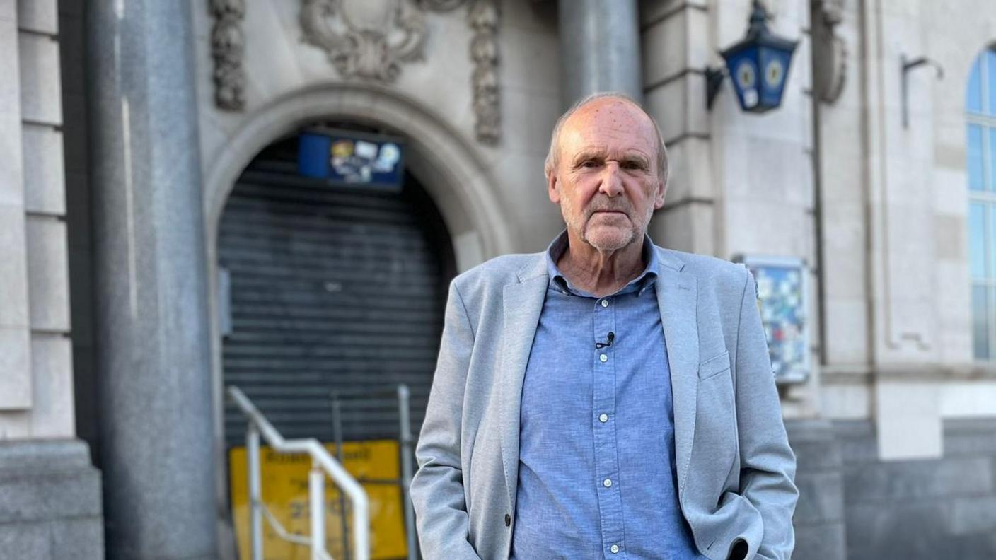 A man in a grey jacket, blue shirt and jeans standing outside a police station. He has thinning grey hair and his hands are in his jean pockets. A police lantern and stone carved insignia can be seen on the grey stone building behind, which has steps up to a shuttered entrance, with marble pillars either side.